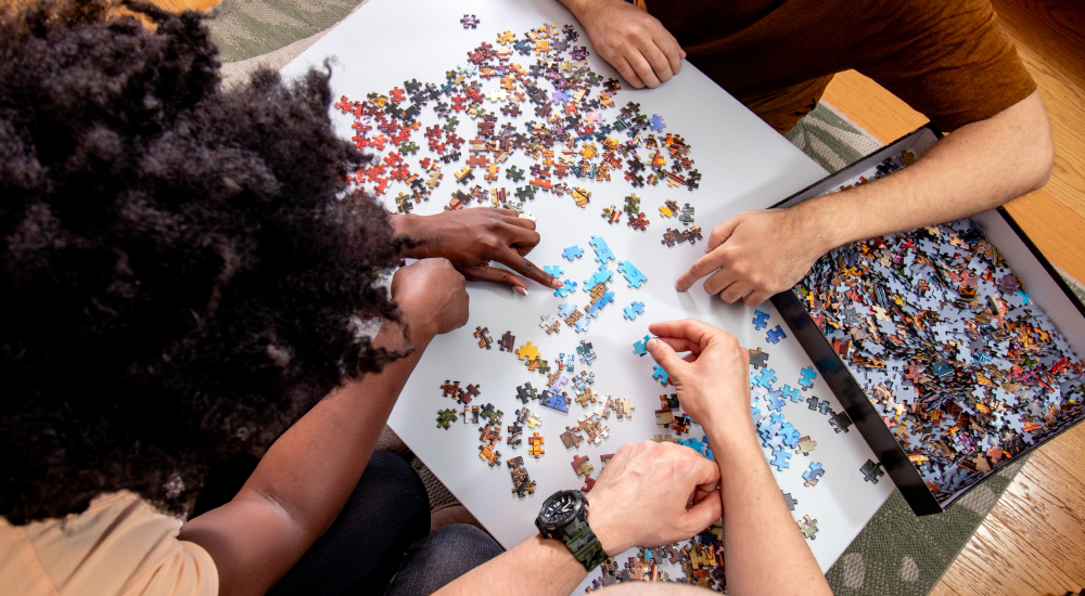 Three people sit around a table, collaborating on a jigsaw puzzle with loose pieces spread out.